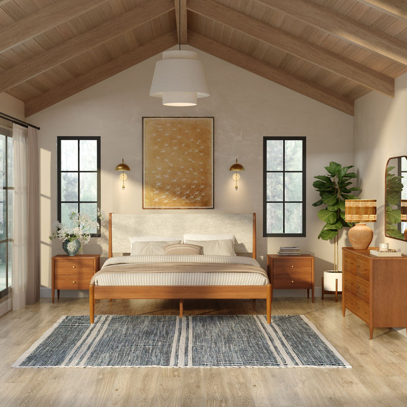 Mid-century modern bedroom with wooden platform bed, vaulted wood-beamed ceiling, large windows, brass wall sconces, matching nightstands, woven area rug and potted plant in a neutral, natural-light interior