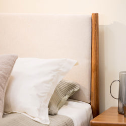 Beige upholstered headboard with wooden post, white pillow and neutral quilted bedding beside a wooden nightstand with glass pitcher
