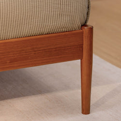 Close-up of solid wood mid-century modern bed frame leg and side rail with beige quilted bedding on a light area rug