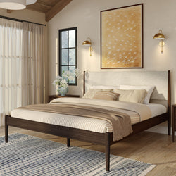 Cozy modern master bedroom with walnut mid-century bed frame, neutral striped bedding and beige throw, large framed wall art, gold wall sconces, natural light and patterned area rug.