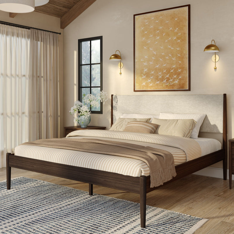 Cozy modern master bedroom with walnut mid-century bed frame, neutral striped bedding and beige throw, large framed wall art, gold wall sconces, natural light and patterned area rug.