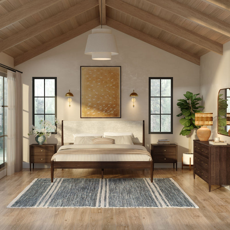 Modern farmhouse bedroom with vaulted wood ceiling, king bed with neutral striped bedding, mid-century wooden nightstands and dresser, blue patterned area rug, large black-framed windows, brass wall sconces, woven lamp and potted fiddle leaf fig plant