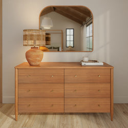 Light oak six-drawer dresser with brass knobs topped by a rattan-woven lamp on a terracotta base and an arched mirror reflecting a neutral boho bedroom — modern Scandinavian farmhouse decor