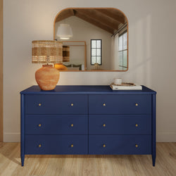 Navy blue six-drawer dresser with brass knobs, woven table lamp and arched mirror in a modern boho bedroom with wood floors
