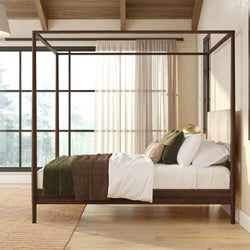 Sunlit modern bedroom with dark wood four-poster bed, white linens, green and brown throw blankets and pillows, sheer curtains and large black-framed windows on light hardwood floor