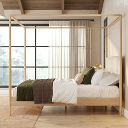 Modern minimalist natural wood four-poster canopy bed in a bright Scandinavian bedroom with white linens, green and brown throws, textured pillows, sheer curtains and large windows letting in natural light