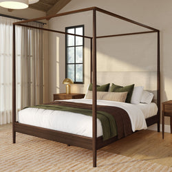 Modern wooden four-poster canopy bed in a neutral bedroom with green and brown bedding, gold bedside lamp, sheer curtains, textured rug and hardwood floors