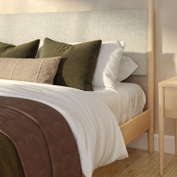 Close-up of a cozy modern bedroom bed with white linen sheets, olive-green textured pillows, beige lumbar cushion, brown throw blanket and light wood bed frame with matching nightstand