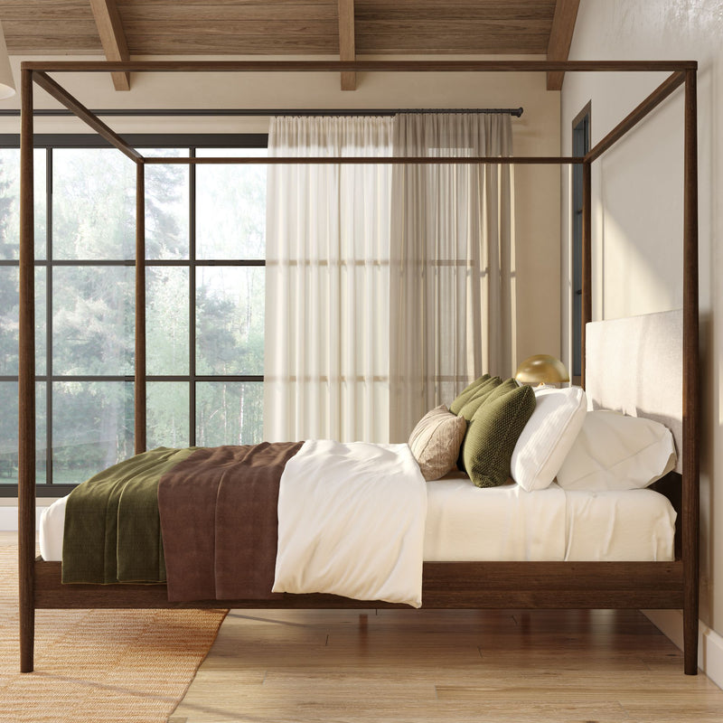 Wooden four‑poster canopy bed in a modern rustic bedroom with neutral linen bedding, green and brown throw blanket and pillows, large floor‑to‑ceiling windows with sheer curtains overlooking a forest, warm natural light, exposed wooden beams and hardwood floor.