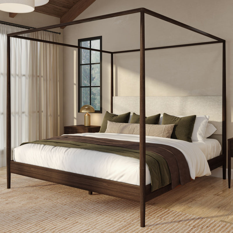 Modern wooden four-poster canopy bed in a neutral bedroom with layered beige and olive bedding, sheer curtains, black-framed window and brass bedside lamp