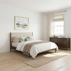 Cozy neutral modern bedroom with wooden platform bed, linen bedding and throw, decorative pillows, textured jute rug, dark wood dresser, framed watercolor art and sunlit window with Roman shade
