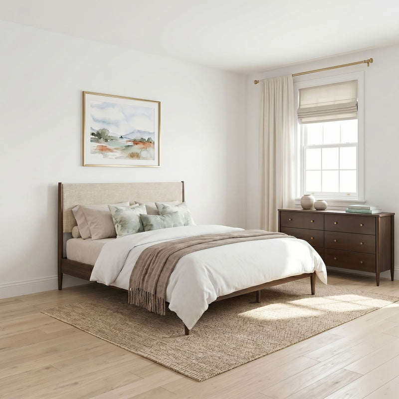 Cozy neutral modern bedroom with wooden platform bed, linen bedding and throw, decorative pillows, textured jute rug, dark wood dresser, framed watercolor art and sunlit window with Roman shade