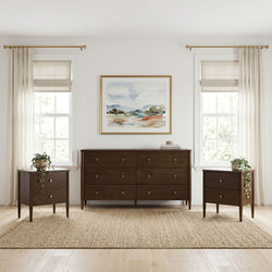 Dark wood mid-century dresser with matching nightstands and potted plants, framed watercolor art between two windows with sheer curtains, beige Roman shades and brass curtain rods, neutral modern bedroom decor