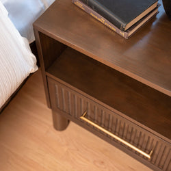 Close-up of a modern walnut nightstand bedside table with brass handle, open shelf and stacked books on a light hardwood bedroom floor