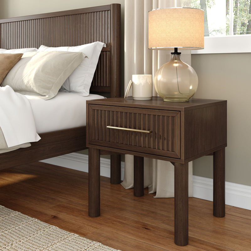 Modern bedroom close-up of dark wood ribbed nightstand with brass-handle drawer, glass table lamp and neutral linen pillows