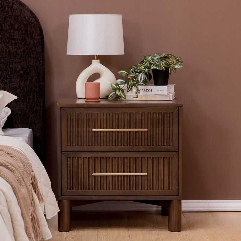 Mid-century modern brown wooden nightstand with two drawers and brass handles topped with a white ceramic lamp, potted plant and books in a styled bedroom