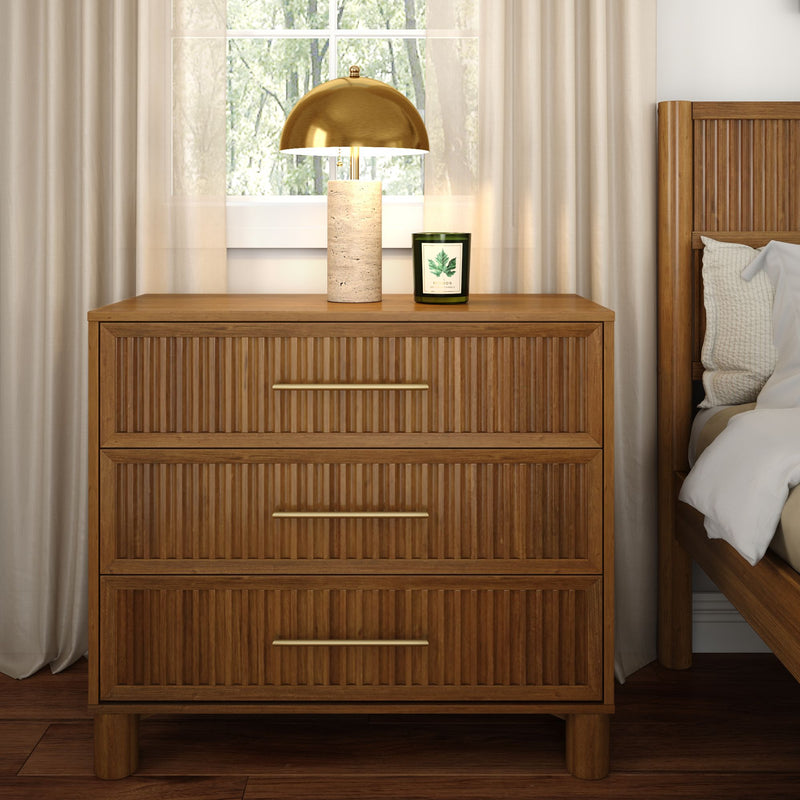 Mid-century modern three-drawer wooden nightstand with brass handles, gold dome table lamp and green scented candle beside a wooden bed in a bright, neutral bedroom with natural light
