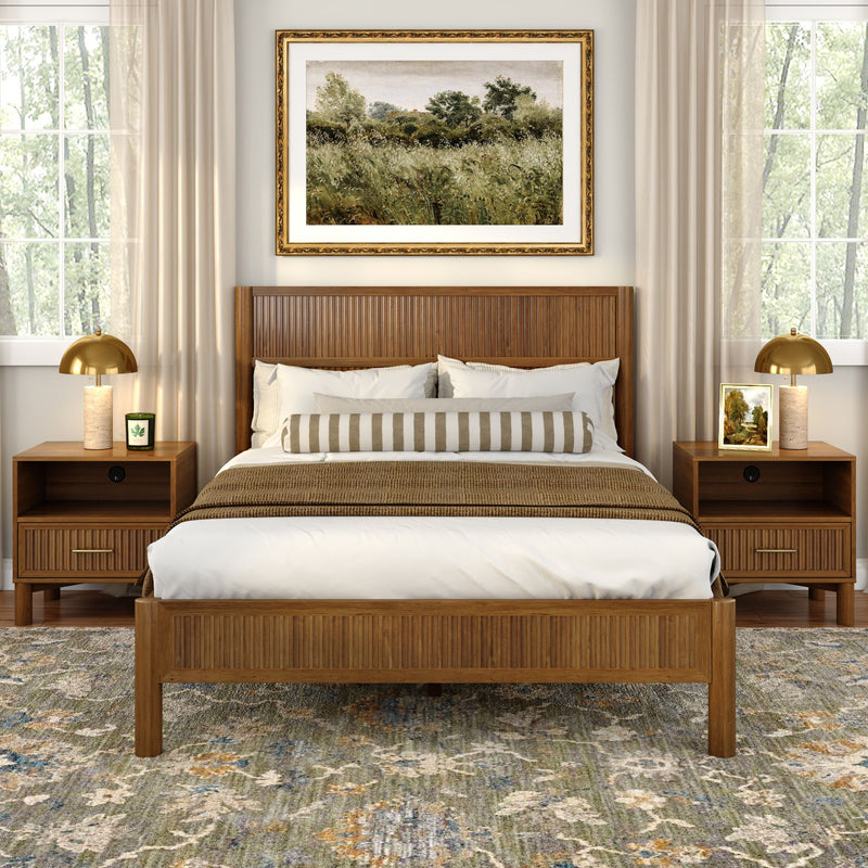 Mid-century modern wood bed with slatted headboard, neutral bedding and striped bolster, matching nightstands with gold lamps, framed landscape art, beige curtains and patterned area rug in a sunlit bedroom