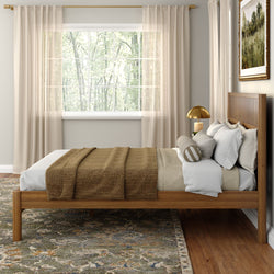 Cozy minimalist bedroom with wooden platform bed, neutral bedding and brown throw, large window with sheer curtains, brass bedside lamp and patterned area rug