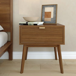 Mid-century modern wooden nightstand with slatted drawer and brass handle, topped with decorative bowl, stacked books and framed abstract art on hardwood floor beside bed