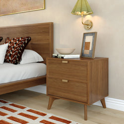 Mid-century modern two-drawer wooden nightstand with brass handles beside bed featuring geometric throw pillow, wall sconce, framed art, books and decorative bowl in a contemporary bedroom