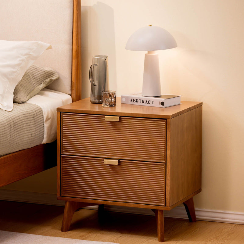 Mid-century wooden nightstand with ribbed drawers and brass handles, white bedside lamp, glass carafe and book — minimalist bedroom decor