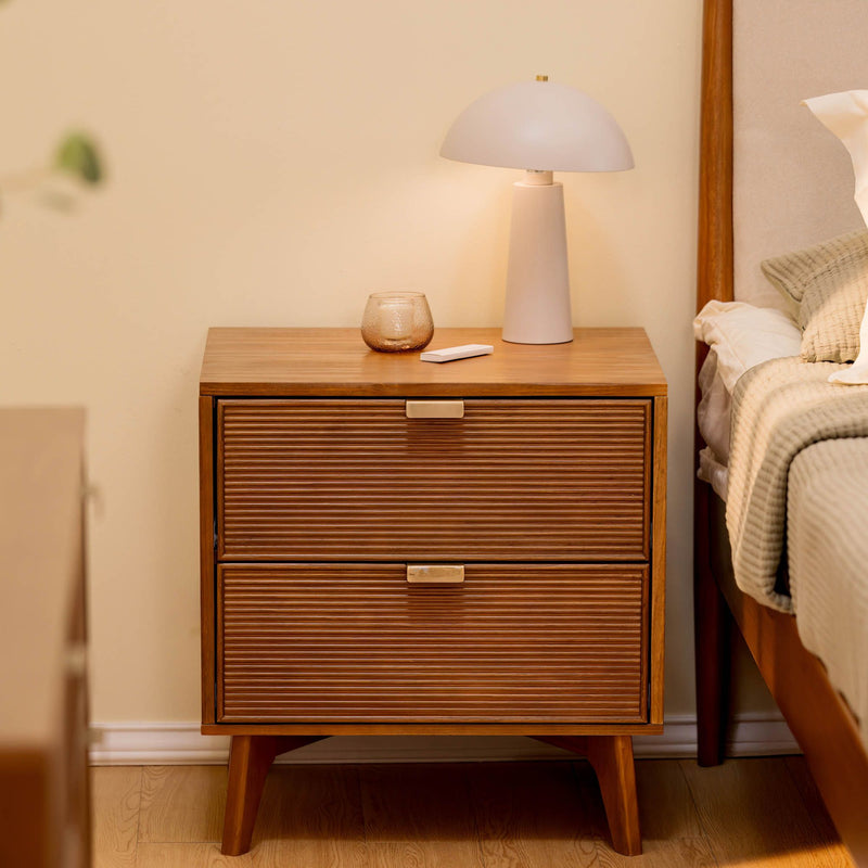 Mid-century modern wooden nightstand with ribbed two-drawer front, white table lamp, glass candle holder and remote beside beige bed