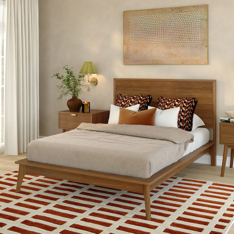 Mid-century modern wooden platform bed in cozy neutral bedroom with geometric accent pillows, red-and-white patterned rug, nightstand, wall art and potted plant