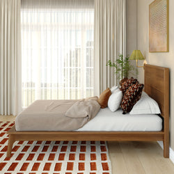 Modern minimalist bedroom with wooden platform bed, neutral linen bedding, patterned throw pillows, geometric red-and-white rug, sheer curtains and natural light