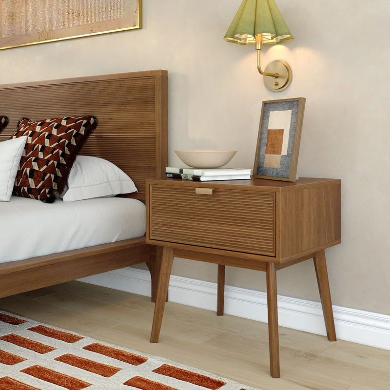 Mid-century modern bedroom with wooden slatted headboard and nightstand, geometric rust-patterned throw pillow on white bedding, brass wall sconce with green shade, framed abstract art, books and decorative bowl, orange-accent area rug