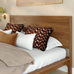 Modern wooden bed with slatted headboard, white sheets and beige linen duvet, rust-and-black geometric throw pillows and brass wall sconce in a cozy contemporary bedroom
