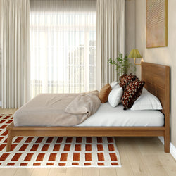 Modern minimalist bedroom interior with wooden platform bed, beige duvet, patterned accent pillows, sheer floor-to-ceiling curtains, potted plant and red brick-pattern area rug