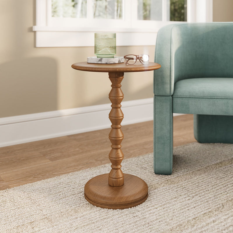 Wooden spindle side table with green ribbed glass candle on marble coaster and eyeglasses beside teal armchair in sunlit living room with hardwood floor and neutral rug
