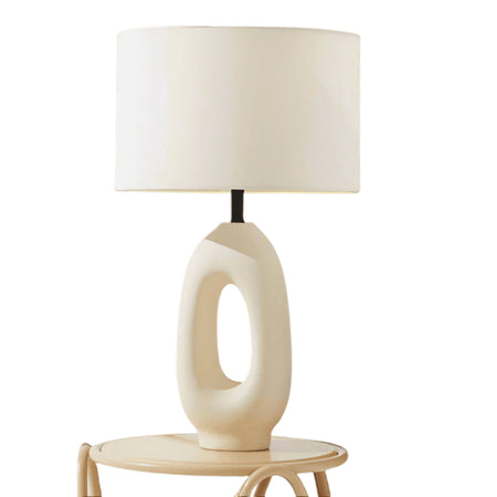 Modern ceramic table lamp with hollow oval base and white drum shade on round side table, minimalist neutral home decor