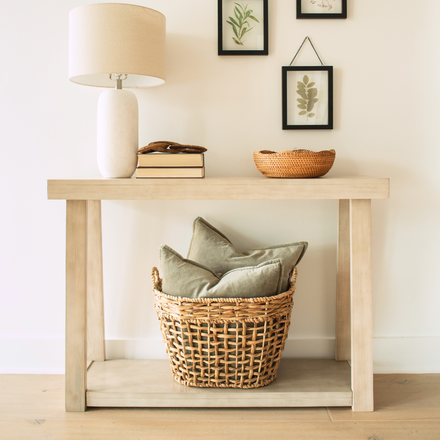 Entryway console table with ceramic lamp, stacked books, woven bowl, framed botanical prints and rattan basket of sage-green pillows