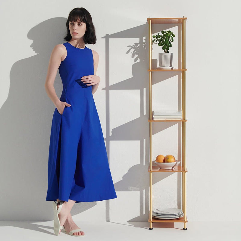 Woman wearing a royal blue sleeveless midi dress and white sandals posing next to a narrow wooden shelving unit with a potted plant, stacked plates and a bowl of oranges, minimalist women's fashion and home decor