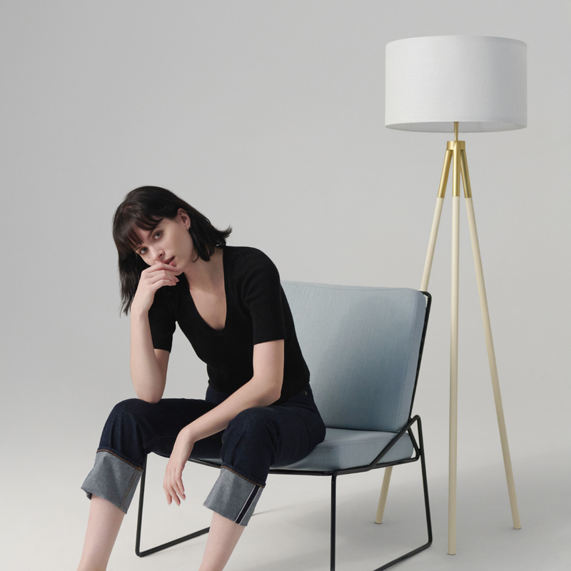Woman seated on light-blue modern lounge chair in minimalist living room, wearing a black top and rolled-cuff jeans beside a white-and-gold tripod floor lamp