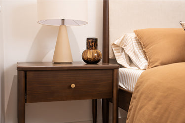 Modern wooden nightstand with beige table lamp and amber glass vase beside bed with tan striped sheets and warm brown duvet