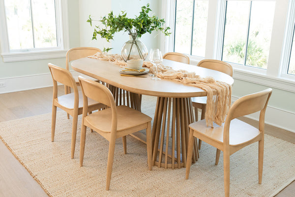 Bright coastal modern dining room featuring an oval light-wood 6-piece dining table set with matching chairs, natural rug, glass vase greenery and large windows