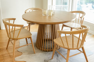 Round oak dining table with four spindle-back wooden chairs and candle centerpiece in sunlit minimalist dining room
