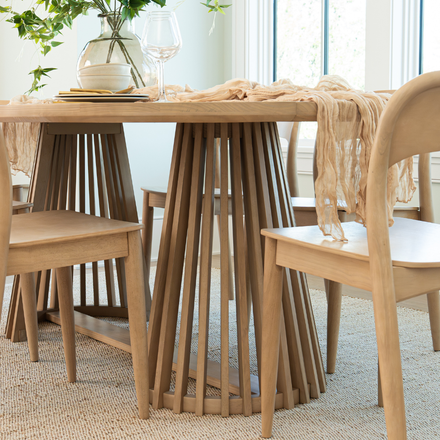 Light oak round dining table with slatted spindle pedestal and matching wooden chairs, styled with glass vase and gauzy table runner in a bright Scandinavian farmhouse dining room