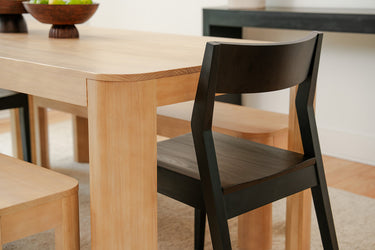 Close-up of light wood dining table corner with black wooden chair and matching bench in a minimalist modern dining room with natural wood finish