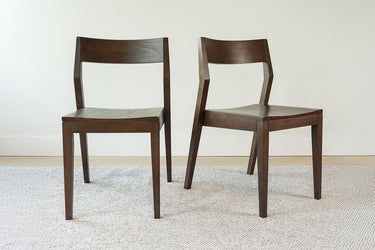 Pair of dark wood dining chairs with angular backs on beige carpet against a white wall