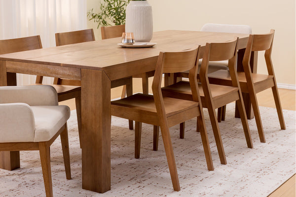 Solid wood dining table with matching wooden dining chairs and an upholstered armchair on a neutral area rug in a bright minimalist dining room, natural wood finish