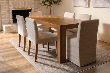 Solid wood rectangular dining table with six beige upholstered chairs on a rug in a neutral dining room with brick fireplace and hardwood floors.