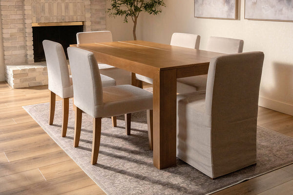 Solid wood rectangular dining table with six beige upholstered chairs on a rug in a neutral dining room with brick fireplace and hardwood floors.