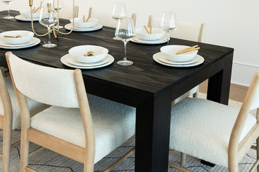 Modern black wood dining table with white stoneware plates and bowls, gold flatware, wine glasses and cream boucle dining chairs in a minimalist luxury dining room