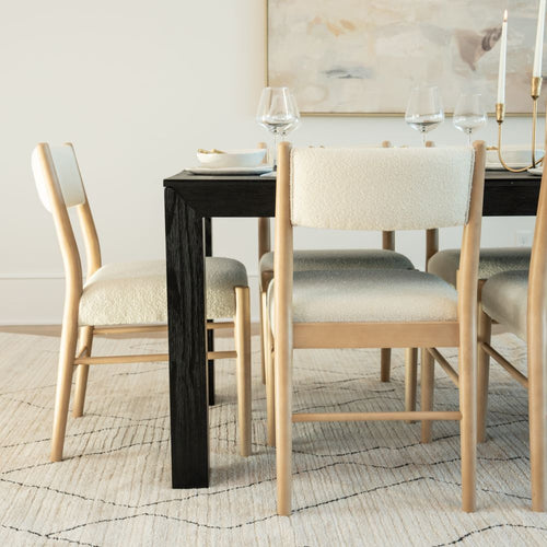 Modern dining room with black wood dining table, light wood cream-upholstered chairs, neutral rug, wine glasses and brass candlesticks.