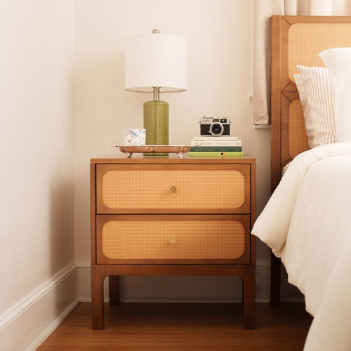 Mid-century modern rattan and wood nightstand with two drawers, green ceramic table lamp, stack of books and vintage film camera next to a white bed in a neutral bedroom
