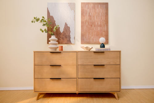 Light oak mid-century modern six-drawer wooden dresser with black handles, styled with sculptural vases, a small globe lamp and abstract copper wall art on a pale yellow wall